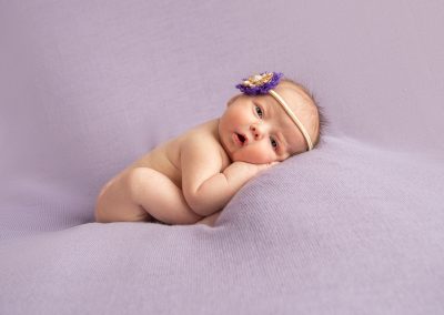 Newborn baby girl on purple backdrop with eyes wide open.